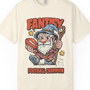 Könnte beinhalten: Cremefarbenes T-Shirt mit einem Cartoon-Zauberer, der einen Football und einen Stab hält. Der Zauberer trägt einen blauen Hut und ein rotes Outfit. Die Worte "FANTASY" und "FOOTBALL CHAMPION" sind orange gedruckt.