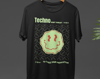 May include: Black t-shirt with a green smiley face design and the text "Techno DON'T FORGET TO TAKE YOUR MEDICATION". The shirt is hanging on a wooden hanger against a dark background.