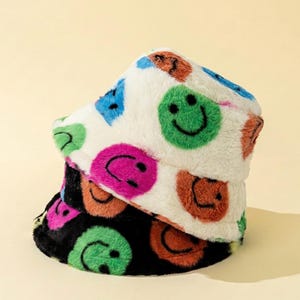 May include: Two fluffy bucket hats stacked on top of each other. The top hat is white with colorful smiley faces in green, pink, blue, and orange. The bottom hat is black with green and orange smiley faces.