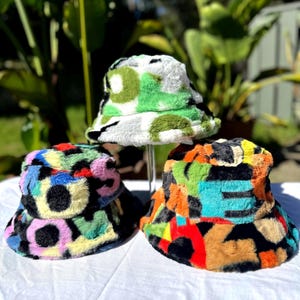 May include: Three fuzzy bucket hats with different colourful abstract patterns. The hats are made of a soft, plush material and have a wide brim.