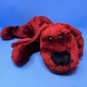 May include: A plush, red dog toy with a black heart-shaped nose and black eyes. The dog is lying down on a blue surface. The toy is made of a soft, fuzzy material.