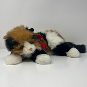 May include: A plush calico cat toy with a red bow. The cat has a mix of brown, black, and white fur, with white paws and a pink nose. The toy is lying down on a white surface.