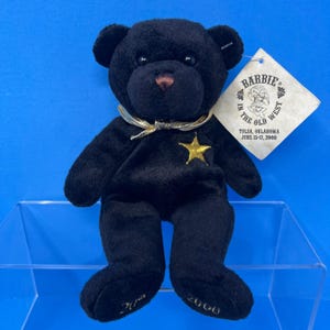 Op de afbeelding: Een zwarte pluche teddybeer met een gouden sterembleem op de borst, een metallic gouden lint om de nek en gouden borduursels op de voeten met de tekst 20 en 2000. Een wit label aan het oor vermeldt BARBIE IN THE OLD WEST TULSA, OKLAHOMA JUNE 15-17, 2000.