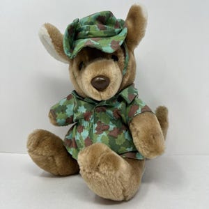 May include: A plush kangaroo toy wearing a camouflage hat and shirt. The kangaroo is brown with a light brown face and ears. The camouflage pattern includes green, brown, and blue-green shapes.