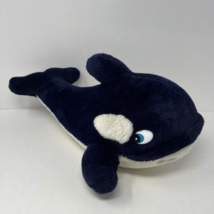 Puede incluir: Un peluche de orca con un cuerpo suave de color azul marino oscuro, vientre blanco y una mancha blanca distintiva cerca del ojo. El juguete de peluche tiene una aleta dorsal prominente, aletas laterales, una cola y un ojo grande.
