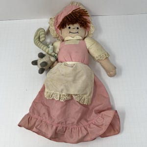 May include: A vintage handmade doll with a pink dress, apron, and bonnet. The doll has brown yarn hair, a smiling face, and holds a striped green and white cane with a small stuffed sheep. The doll's dress has a ruffled hem.