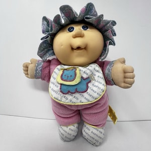 Puede incluir: Una muñeca Cabbage Patch Kids con cara redonda, ojos azules y hoyuelos. La muñeca lleva un gorro con volantes de patrón gris y azul, un conjunto de forro polar rosa y un babero blanco con el texto BABYLAND KID y un gráfico de león azul.