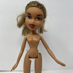 May include: A Bratz doll with tan skin, brown eyes, and brown hair. The doll has full lips and is wearing makeup. The doll is nude and has articulated arms and legs. The doll is standing upright.