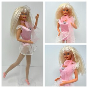 May include: A blonde doll with blue eyes wearing a pink ribbed leotard, a sheer white skirt with silver star patterns, a matching pink scarf, and pink ballet slippers. The doll has long straight hair with a small braided section on one side.
