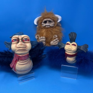 Pode incluir: Três figuras de criaturas fantásticas sobre um fundo azul. Uma é marrom e peluda com chifres, e duas têm pele clara, olhos vermelhos e detalhes de penas azuis. Uma tem um cachecol vermelho.