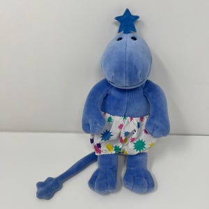 Puede incluir: Un peluche azul con forma de criatura con una estrella en la cabeza. Tiene una larga cola y lleva pantalones cortos blancos con patrones de estrellas de colores. Juguete suave y tierno para niños.