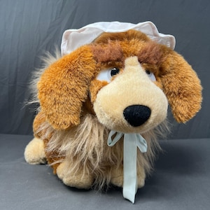 May include: A plush dog toy with brown fur, a tan muzzle, and a black nose. The stuffed animal wears a white bonnet tied under its chin with a light blue ribbon. It has large white eyes with black pupils and long, shaggy brown ears.