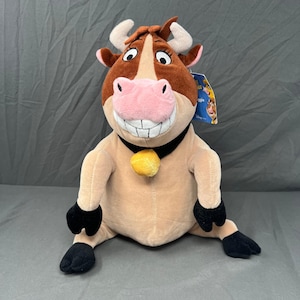 May include: A plush cow toy featuring a brown and tan body, a large pink snout, and a wide grin with visible teeth. The cow wears a black collar with a yellow bell and has small white horns. A blue product tag is attached to the side of the head.