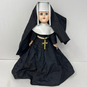 May include: A doll dressed in a traditional black and white nun habit, featuring a long black gown, a white wimple, and a black veil. The doll wears a white cord belt around the waist with a gold-coloured crucifix pendant hanging from the centre.
