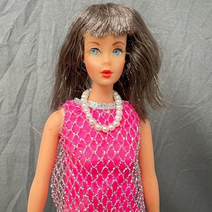 May include: A doll with shoulder-length brown hair, blue eyes, and red lips. The doll wears a sleeveless top made of bright pink fabric with a silver metallic diamond-shaped mesh overlay and a white faux pearl necklace around the neck.