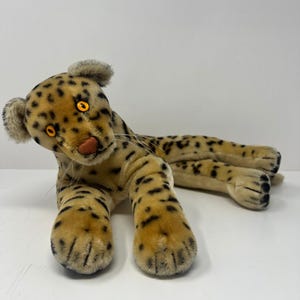 May include: A plush leopard toy with a tan body, black spots, and a cream-colored underbelly. The leopard has yellow eyes, a pink nose, and long whiskers. The toy is lying down with its head turned to the side.