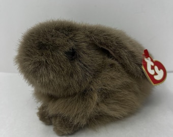 May include: A small, brown, plush rabbit toy with soft, fuzzy fur and long, floppy ears. The stuffed animal features a dark, round eye and a small, red, heart-shaped Ty tag attached to the side, displaying the white text ty in a stylized font.