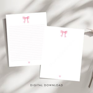 May include: Two sheets of white stationery paper featuring a watercolor-style pink bow at the top center and a small pink heart at the bottom center. One sheet contains horizontal lined writing space, while the other sheet is blank.
