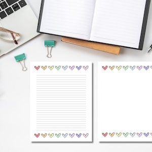 Pode incluir: Duas folhas de papelaria brancas com uma borda de coração em várias cores. Uma folha é pautada e a outra é em branco. A papelaria está em uma superfície branca com material de escritório, incluindo um laptop e clipes.