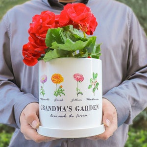 May include: White ceramic planter with a floral design and the text "Grandma's Garden love each other forever". The planter has four names printed on it: Nicole, Sarah, Jillian, and Madison. A person is holding the planter with red flowers.