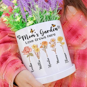 May include: White ceramic Mom's Garden planter with the text "Mom's Garden Love Grows Here" and illustrations of flowers. The planter includes a matching saucer and is filled with purple and green artificial flowers.
