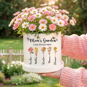May include: A white ceramic flower pot filled with pink and white daisies. The pot is decorated with the words "Mom's Garden" and "Love Grows Here", along with floral illustrations and names. Two butterflies are also depicted on the pot.