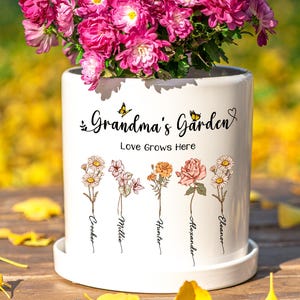 May include: A white ceramic plant pot with the text "Grandma's Garden" and "Love Grows Here". The pot has floral illustrations and names. It is filled with bright pink flowers and sits on a white saucer, perfect for a garden.