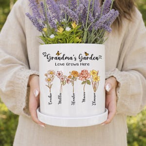 以下が含まれることがあります： 「Grandma's Garden」と「Love Grows Here」の黒い文字が書かれた、白い円筒形のセラミック製プランターと受け皿。Crocker、Millie、Hunter、Maeve、Eleanorという名前が添えられた、5つのカラフルな花のイラストが描かれています。