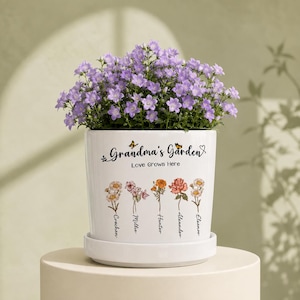 Peut inclure: Un pot de fleurs en céramique blanche avec sa soucoupe, rempli de campanules violettes. Le pot porte l'inscription "Grandma's Garden Love Grows Here" avec des papillons et cinq tiges de fleurs illustrées portant les noms Coachen, Millie, Hunter, Alexander et Eleanor.