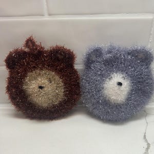 May include: Two knitted dishcloths shaped like bear faces. One is brown with a beige face and the other is purple with a white face.