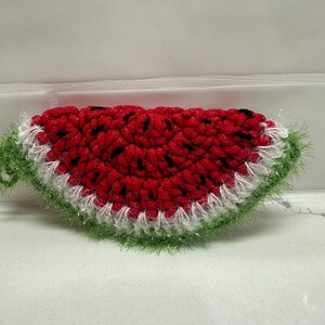 May include: A crocheted watermelon slice with red, black, white, and green yarn. The red center has black seed details, a white border, and green rind with fuzzy yarn. This handmade item is a decorative piece.