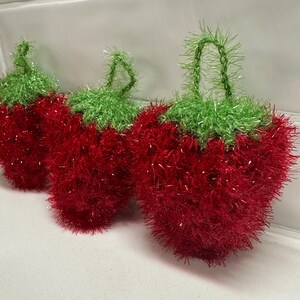 May include: Three red and green crocheted strawberry-shaped dishcloths. The dishcloths are made with a sparkly yarn and have a loop at the top for hanging.