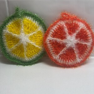 May include: Two knitted dishcloths shaped like citrus fruit slices. One is green and yellow, resembling a lime, and the other is orange and white, resembling an orange.