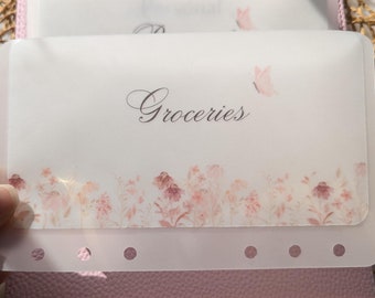 May include: A clear plastic budget envelope with the word "Groceries" in elegant script. The envelope features a floral design in shades of pink and a small butterfly. The envelope is held over a pink planner.