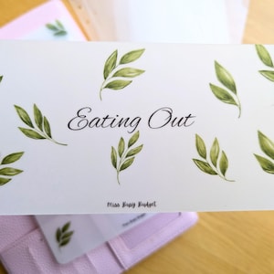 May include: A white card featuring the words "Eating Out" in a stylish font, decorated with green watercolour leaf designs. The card is held above a pink wallet, possibly for budgeting or organisation.