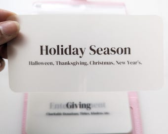 May include: A white card with the text "Holiday Season" and "Halloween, Thanksgiving, Christmas, New Year's". The card is held in a hand over a pink leather planner.