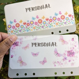 Op de afbeelding: Twee witte plastic scheiders met een bloemenontwerp en het woord "PERSOONLIJK" erop gedrukt. De bovenste scheider heeft een kleurrijk bloemenontwerp met vlinders. De onderste scheider heeft een aquarel bloemenontwerp met vlinders in roze en paarse tinten.