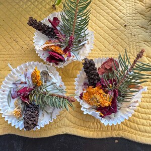 May include: Three white paper cupcake cases filled with dried flowers, pine needles, and small pine cones. The flowers are orange, red, and yellow. The cases sit on a yellow quilted surface.