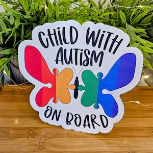 Könnte beinhalten: Ein weißer Aufkleber mit dem Text "CHILD WITH AUTISM ON BOARD" in Schwarz. Der Aufkleber zeigt zwei Schmetterlingsdesigns mit regenbogenfarbenen Flügeln. Der Aufkleber befindet sich auf einer Holzoberfläche mit grünem Laub im Hintergrund.