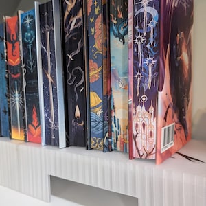 May include: A row of hardcover books standing upright on a white shelf. Each book features a unique, colorful spine with intricate illustrations including swords, stars, flames, candles, and architectural designs. The books are arranged side by side.