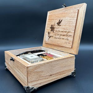 May include: Wooden jewelry box with a hinged lid, featuring a laser-etched design of a bird and flower, and an inspirational quote. The box contains various items, including sunglasses and small containers. The box has decorative metal feet.