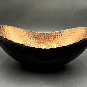 May include: A black hammered metal bowl with a textured exterior and a gold-coloured interior. The bowl has a scalloped rim and an oval shape. The interior has a hammered texture, and the exterior is a matte black finish.