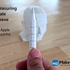 May include: A white measuring scale sleeve for the Apple Pencil Pro, held in front of a white geometric head sculpture. The sleeve has black markings for measurement. The text "Measuring Scale Sleeve for Apple Pencil Pro" is visible.