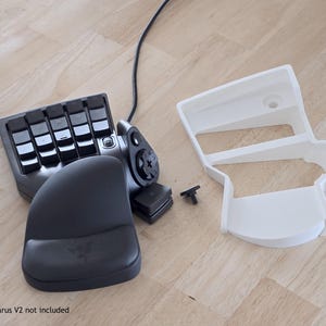 May include: A black gaming keypad with a wrist rest, a white plastic component, and a small black piece. The keypad has multiple keys and a joystick. The Razer logo is visible on the wrist rest.
