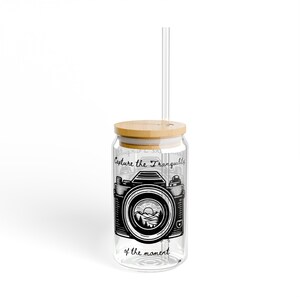 May include: A clear glass tumbler with a bamboo lid and a glass straw. The tumbler has a black and white illustration of a vintage camera with a mountain scene in the viewfinder. The text "Capture the Tranquility of the moment" is printed on the tumbler.