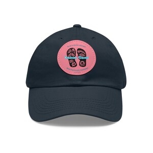 Op de afbeelding: Een donkerblauwe baseballpet met een roze patch met daarop een paar slippers met bloemenmotieven. De patch heeft de tekst "Business Casual" en "Black Dog Murphy Beachwear".