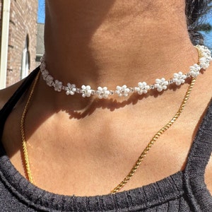 May include: A white beaded choker necklace with small flower-shaped beads. The necklace is worn on a person's neck.