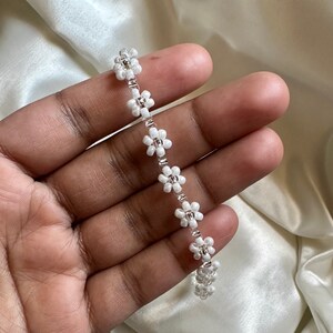 May include: A white beaded choker necklace with small flower-shaped beads. The necklace is on a hand.