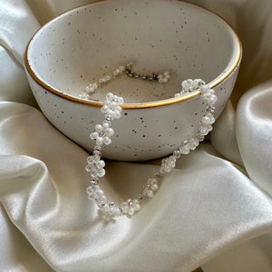 May include: A white beaded choker necklace with small flower-shaped beads. The necklace is lying in a white ceramic bowl with a speckled glaze.