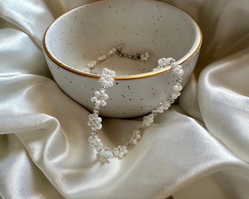 May include: A white beaded choker necklace with small flower-shaped beads. The necklace is lying in a white ceramic bowl with a speckled glaze.